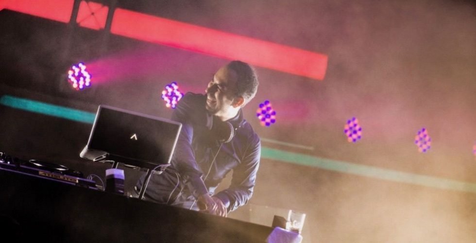 A smiling DJ performs at a night event, wearing headphones and using a laptop on a stage illuminated by vibrant pink spotlights and a red neon light bar through hazy smoke.