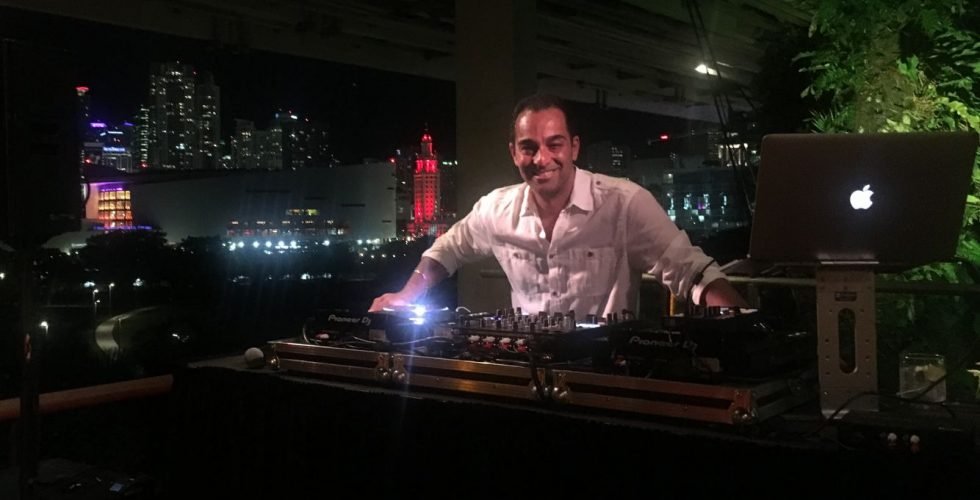 A smiling DJ performs at a night event, wearing headphones and using a laptop on a stage illuminated by vibrant pink spotlights and a red neon light bar through hazy smoke.