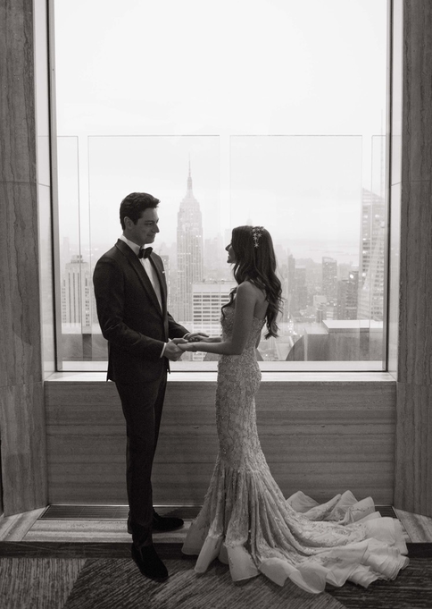 Black and white photo of a bride in a mermaid-style gown and a groom in a tuxedo holding hands in front of a large window with a view of the New York City skyline.