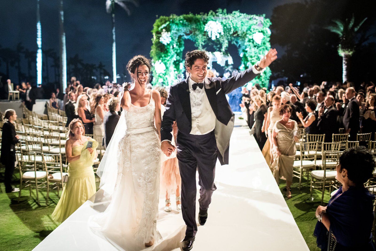 Alexa and Nico at their destination wedding in West Palm Beach, FL A bride and groom walking down the aisle at an outdoor night wedding ceremony surrounded by guests and palm trees.